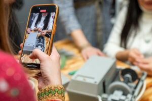 A person recording a video of a sewing machine with their phone, showing hands working with thread, for the purpose of social media storytelling.