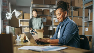 A woman in a warehouse using a laptop to apply for small business grants, with a man packing a box in the background.